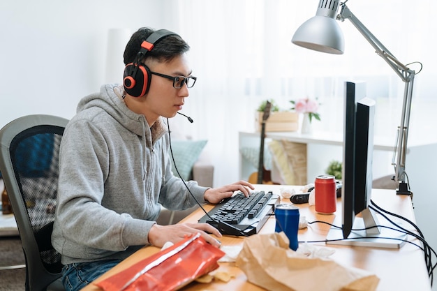 Technologia Gier Rozrywka Koncepcja. Szczęśliwy Młody Człowiek Azjatyckich W Słuchawkach Z Komputerem Pc, Grając W Gry I Strumieniowe Odtwarzanie Wideo. Leniwy Chłopak Z College'u Odpoczywa W Domu W Przerwie Letniej, Jedz Niezdrowe Jedzenie