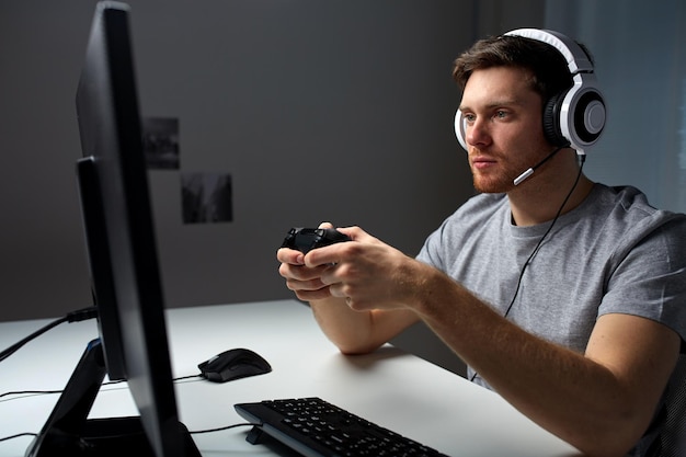 technologia, gry, rozrywka, zagrajmy i koncepcja ludzi - młody człowiek w zestawie słuchawkowym z gamepadem kontrolera grający w grę komputerową w domu i przesyłający strumieniowo grę lub wideo instruktażowe