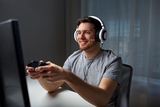 technologia, gry, rozrywka, zagrajmy i koncepcja ludzi - szczęśliwy młody człowiek w zestawie słuchawkowym z gamepadem kontrolera grający w grę komputerową w domu i przesyłający strumieniowo grę lub solucję vid