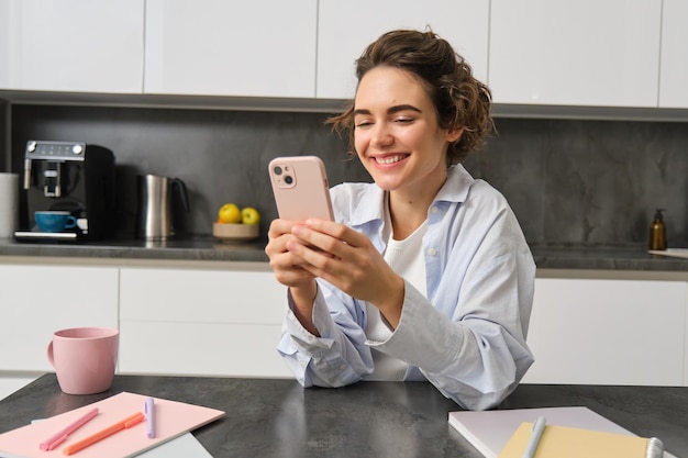 Technologia i styl życia Młoda kobieta siedzi w domu używa smartfona w kuchni i uśmiecha się