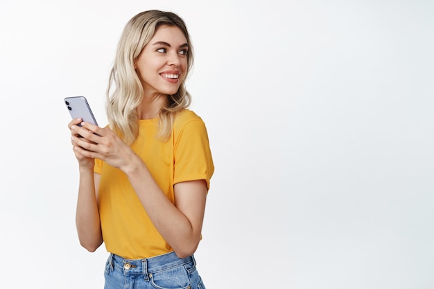 Technologia komórkowa. Młoda blond kobieta trzyma telefon komórkowy, odwraca głowę i uśmiecha się do miejsca kopiowania, stoi w żółtej koszulce na białym