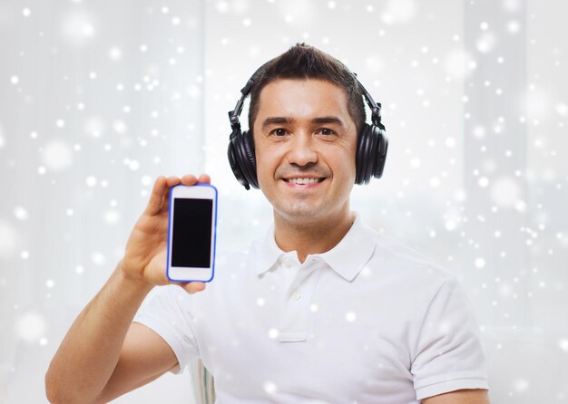 technologia, ludzie, styl życia i koncepcja uczenia się na odległość - szczęśliwy człowiek w słuchawkach pokazujący czarny pusty ekran smartfona i słuchający muzyki w domu przez efekt śniegu