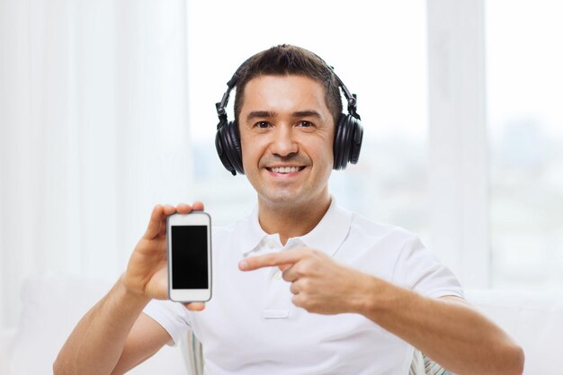 technologia, ludzie, styl życia i koncepcja uczenia się na odległość - szczęśliwy człowiek w słuchawkach pokazujący czarny pusty ekran smartfona i słuchający muzyki w domu