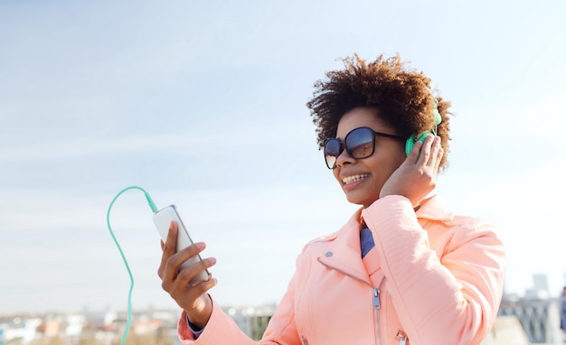 technologia, styl życia i koncepcja ludzi - uśmiechnięta młoda kobieta z Afryki lub nastoletnia dziewczyna ze smartfonem i słuchawkami słuchająca muzyki na świeżym powietrzu