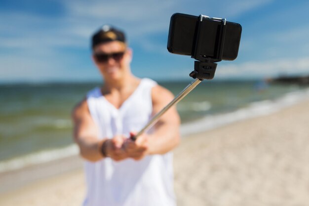 technologia, wakacje letnie i ludzie koncepcja - szczęśliwy młody człowiek robiący zdjęcie za pomocą smartfona na selfie stick na plaży