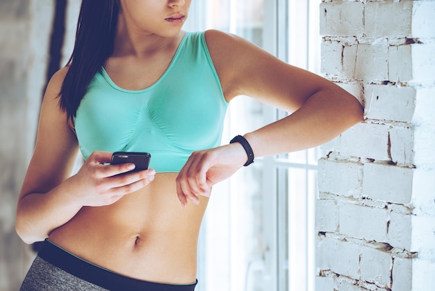 Zdjęcie technologie ułatwiają szkolenie. zbliżenie na część młodej kobiety, która patrzy na swoją sportową bransoletkę i używa smartfona, opierając się o ścianę na siłowni