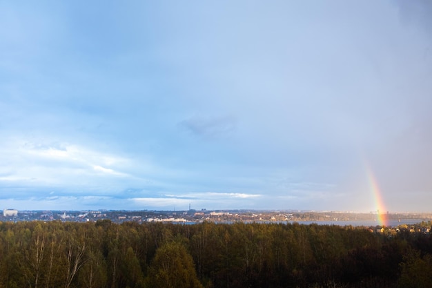 Tęcza po deszczu nad rzeką na tle miasta. Błękitne niebo, żółte drzewa