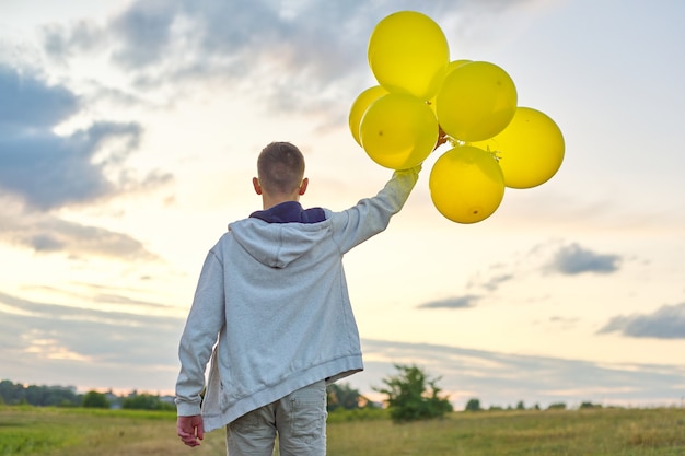 Teen chłopiec biegnący z balonami, widok z tyłu. Natura, łąka, niebo w tle chmur. Wakacje, urodziny, koncepcja wolności stylu życia