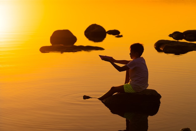 Teen chłopiec ze statkiem origami w ręce. Chłopiec siedzi na skale w pobliżu rzeki. Piękny pomarańczowy zachód słońca. Origami. Rzeka. Jezioro. Letnie powołanie