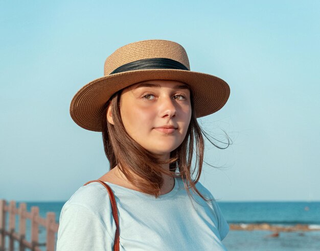 Teen dziewczyna stojąca na drewnianym chodniku nad morzem o zachodzie słońca, ubrana w jasnoniebieski t-shirt i ha słomy. Letnie podróże i blisko koncepcji natury.