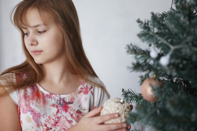 Teen dziewczyna w pobliżu choinki. Nastolatek z długimi włosami w kolorowej sukience zdobi zabawki sylwestrowe.