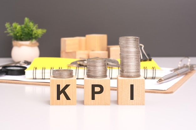 Tekst KPI w drewnianych blokach z monetami ułożonymi w rosnące stosy Koncepcja kluczowych wskaźników wydajności