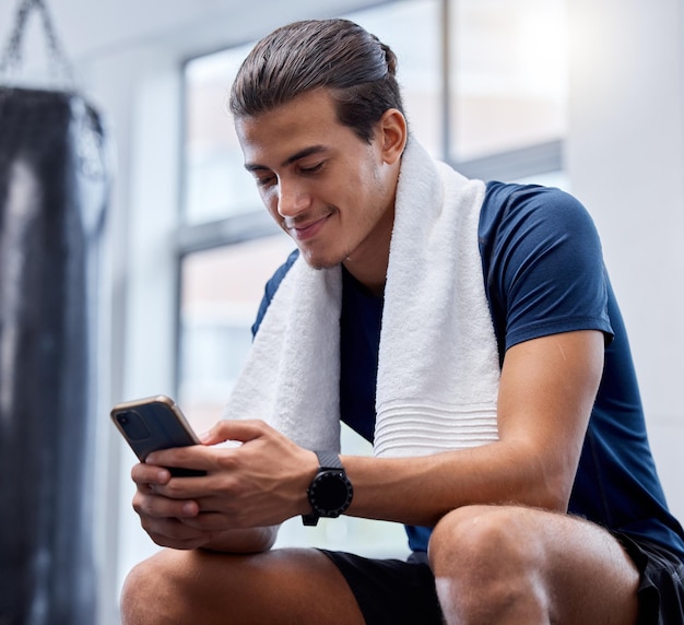 Telefon fitness i mężczyzna relaksują się na siłowni, śledząc ćwiczenia lub postępy w aplikacji Sporty techniczne i szczęśliwy mężczyzna z ręcznikiem i smartfonem mobilnym do przeglądania internetu, wiadomości tekstowych lub mediów społecznościowych