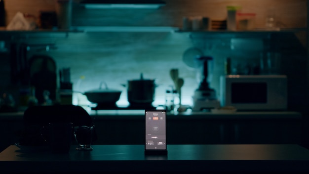 Telefon z inteligentnym oprogramowaniem umieszczony na stole w kuchni, gdzie nikogo nie ma, kontrolujący światło za pomocą zaawansowanej technologii. Mobilny z aplikacją smart home w systemie automatyki pustego domu