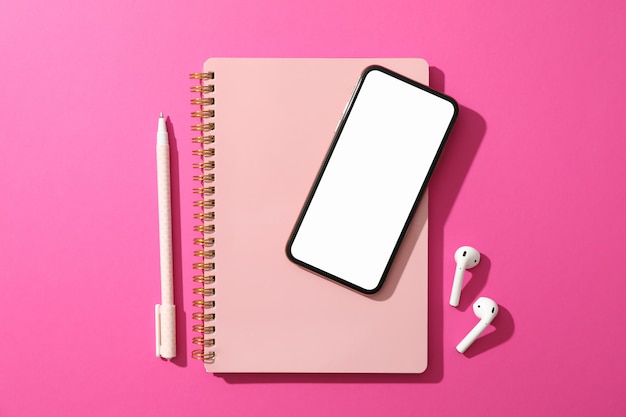 Telefon z pustym ekranem, słuchawkami, zeszytem i długopisem na różowej powierzchni