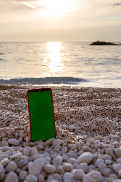 Telefon z zielonym ekranem na skalistej plaży na tle zachodu słońca