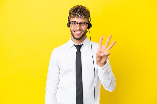 Telemarketer blond mężczyzna pracujący z zestawem słuchawkowym na żółtym tle szczęśliwy i liczący trzy palcami