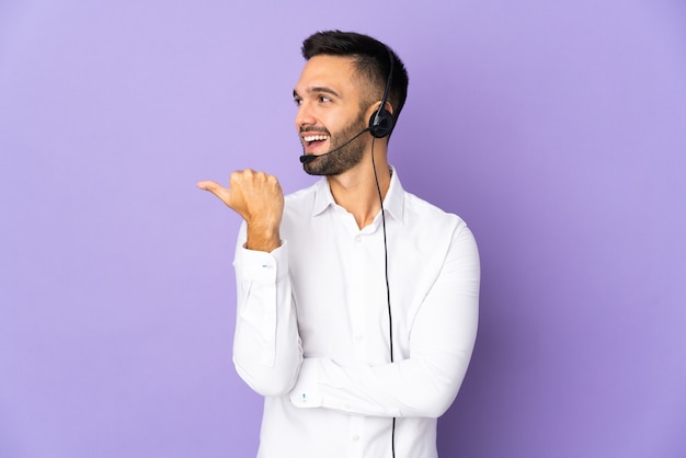 Telemarketer mężczyzna pracujący z zestawem słuchawkowym na białym tle na fioletowym tle, wskazując w bok, aby przedstawić produkt