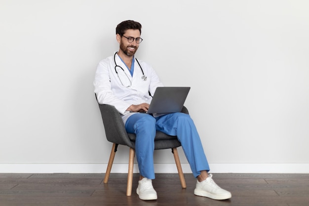 Telemedycyna przystojny mężczyzna lekarz w mundurze siedzi na krześle i za pomocą laptopa