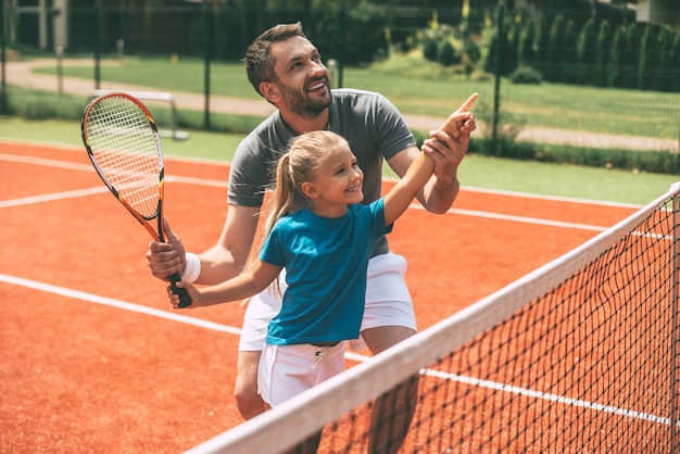 Tenis jest fajny, gdy ojciec jest w pobliżu. Wesoły ojciec w sportowej odzieży uczy córkę grać w tenisa, gdy oboje stoją na korcie tenisowym