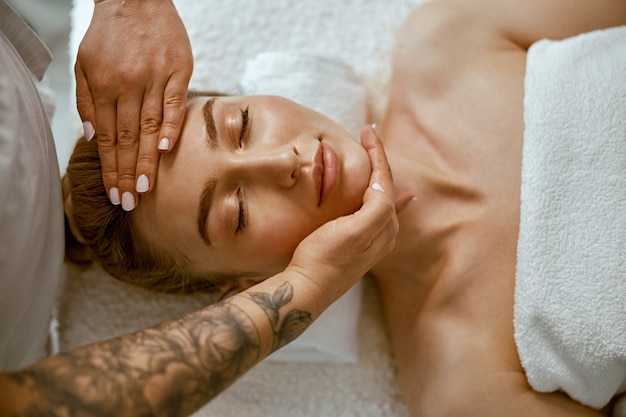 Terapeuta wykonuje masaż relaksacyjny twarzy młodej kobiecie w nowoczesnym gabinecie wellness