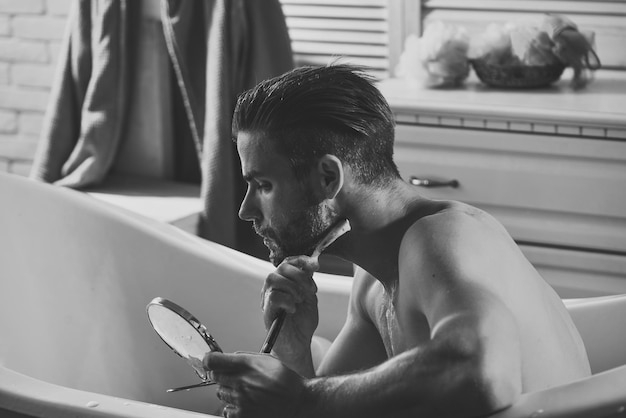 Terapia pielęgnacyjna Brodaty mężczyzna golenie brzytwą w wannie w łazience Golenie fryzjera fryzjer Macho z mydłem do golenia na brodzie włosy spojrzenie w lustrze Pielęgnacja higiena zdrowie