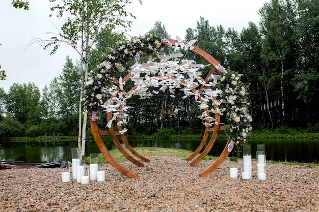 Teren ceremonii zaślubin w lesie, nad rzeką na molo. Drewniany łuk okrągły ozdobiony kwiatami, zielenią, białymi ptaszkami, świecami, krzesłami ławka dla gości. Ładny, stylowy wystrój