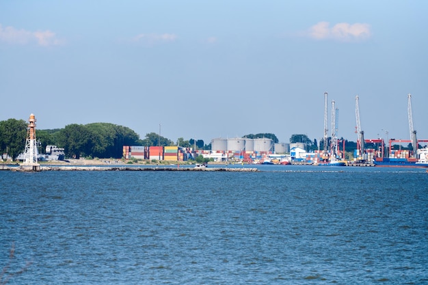 Terminal kontenerowy w logistyce eksportu i importu. Wysyłka ładunku do portu dźwigiem, błękitne niebo i morze, dzień. Transport towarowy, wysyłka na cały świat, logistyka, komunikacja globalna, przemysł