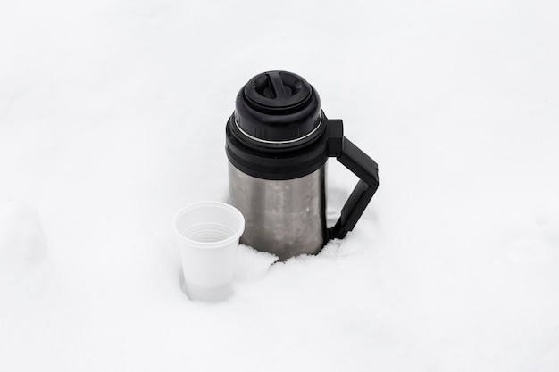 Termos z gorącą herbatą i jednorazową szklanką zimą w zaspie śnieżnej w przyrodzie.