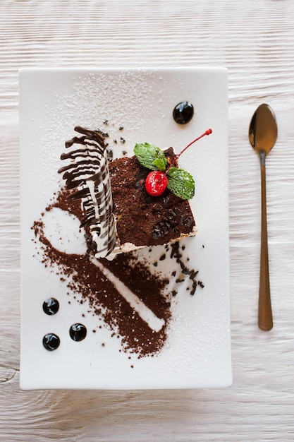 Tiramisu na białym kwadratowym talerzu z dekoracją z czekoladowej wiśni i mięty Słodki deser podawany w restauracji na drewnianym stole widok