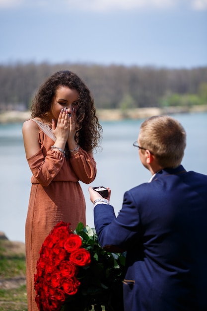 Tło weselne w lesie: mężczyzna zrobił niespodziankę, pierścionek wręcza pięknej dziewczynie deklarację miłości i oświadczyny. Dziewczyna z uśmiechem na twarzy i bukietem czerwonych róż