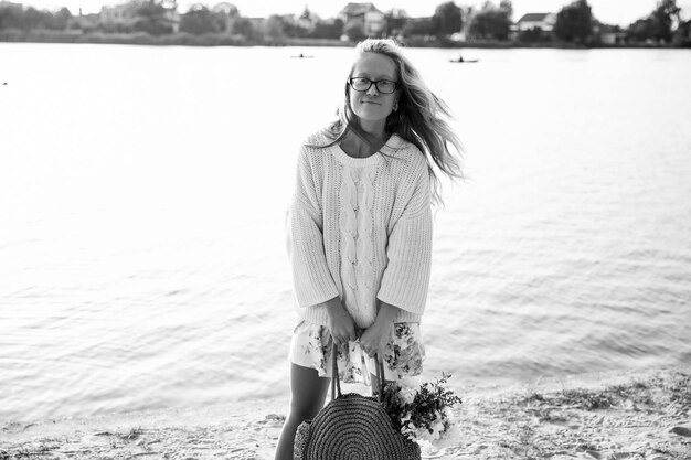 To czarno-białe zdjęcie stockowe pokazuje młodą blondynkę spacerującą po plaży w sukience i swetrze z kwiatami w słoneczny dzień