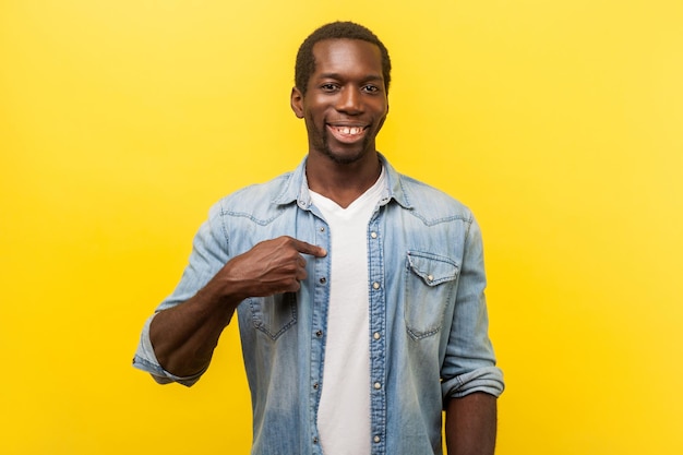 To ja. Portret udanego pozytywnego młodego człowieka w dżinsowej koszuli dorywczo, wskazując na siebie, uśmiechając się szczęśliwie do kamery, ciesząc się z osiągnięć. strzał w studio na białym tle na żółtym tle
