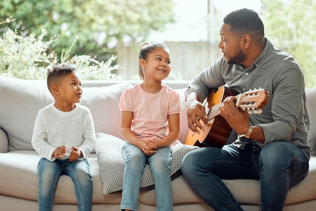 To ulubiona piosenka mamy Ujęcie młodego ojca, który uczy swoje dzieci gry na gitarze, siedząc na kanapie w domu