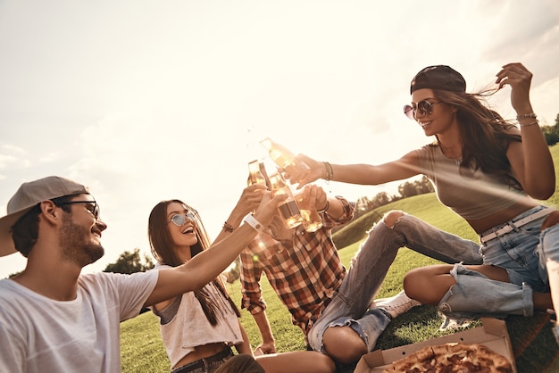 Toast za przyjaciół! Grupa młodych uśmiechniętych ludzi w swobodnym stroju toastów podczas pikniku w ciepły letni dzień na świeżym powietrzu