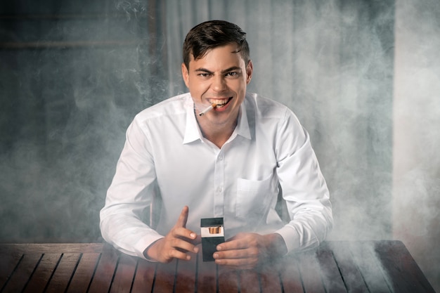 Tobacco Evil: Portret Młodego Faceta O Złym Spojrzeniu Na Zadymionym Tle, Trzymającego W Rękach Paczkę Papierosów I Zapalonego Cygara W Ustach. Złe Nawyki Są Zabijane.