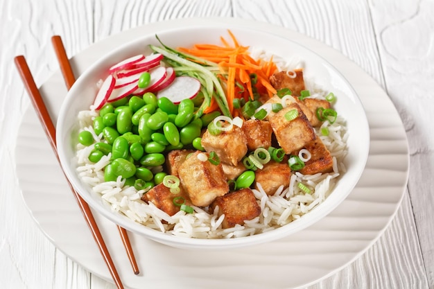 Tofu poke miska z ryżem basmati fasolą edamame rzodkiewką cienko pokrojoną w plasterki ogórka i marchewki w białej misce z pałeczkami widok poziomy z góry