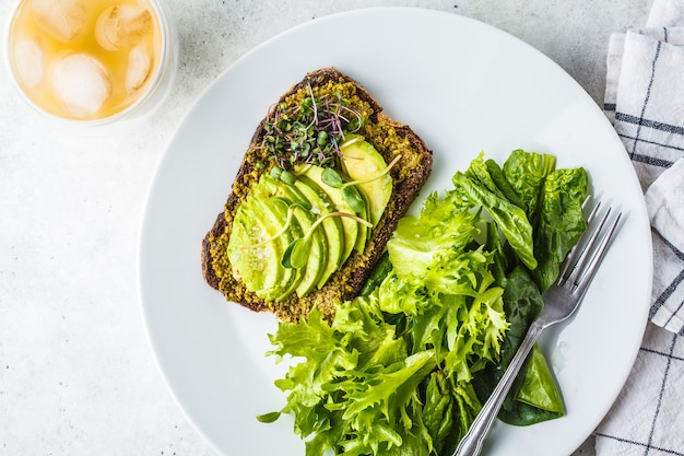 Tost z awokado z pesto, kiełkami i surówką na białym talerzu i mrożoną kawą.