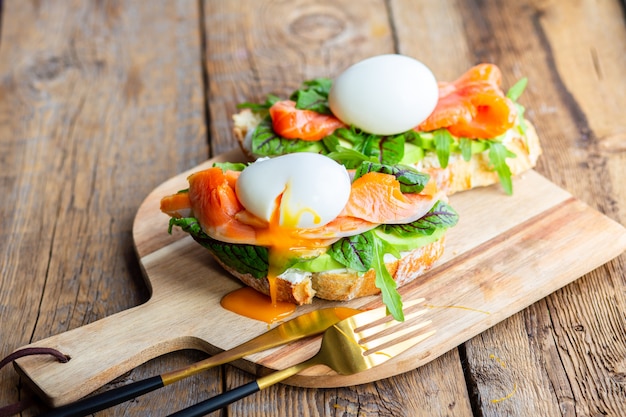 Tosty z jajkiem w koszulce, awokado, rukolą, serem, czerwoną rybą z ziołami. Smaczny lunch. Prawidłowe odżywianie ze zdrowymi tłuszczami. Pyszny tost z awokado. Jajko sadzone rozprzestrzenia się na grzance