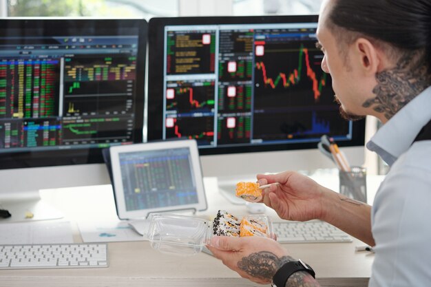 Trader jedzący sushi na lunch i monitorujący dane giełdowe na wielu ekranach komputerów w swoim biurze