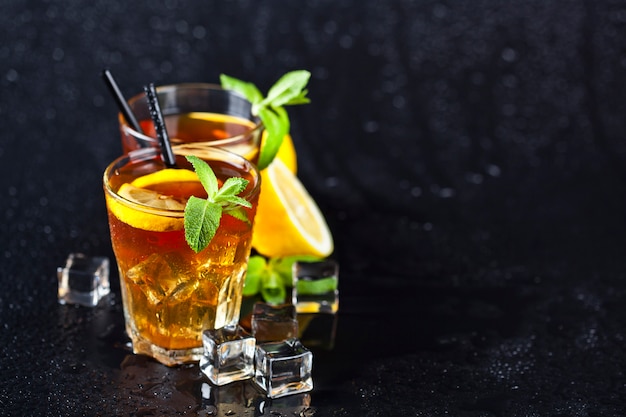 Tradycyjna mrożona herbata z cytryną, liśćmi mięty i kostkami lodu w dwóch szklankach na mokrym czarnym tle.
