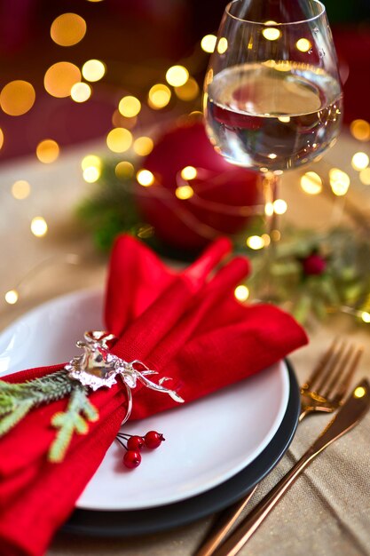 Tradycyjne nakrycie stołu Bożego Narodzenia z bliska. Złote sztućce, lniana serwetka z pierścieniem jelenia, świerkowymi gałązkami i kieliszkiem wina. Tło wakacje. Selektywne skupienie, girlanda, bokeh.