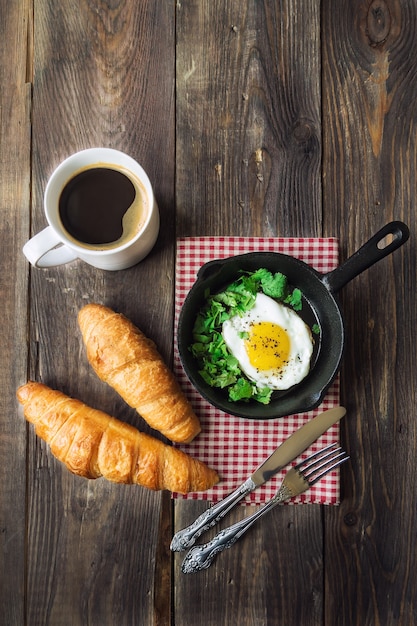 Tradycyjne śniadanie z kawą, rogalikami i jajkiem sadzonym na żelaznej patelni na rustykalnym drewnianym stole.