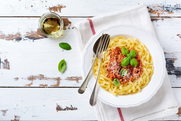 Tradycyjne włoskie danie makaron fettuccine z sosem bolońskim, bazylią i parmezanem w białym talerzu na jasnym drewnianym tle