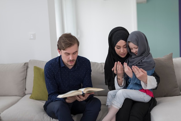 Tradycyjni muzułmańscy rodzice rodzinni z dziećmi czytającymi Koran i modlącymi się razem na kanapie przed kolacją iftar podczas uczty ramadanu w domu