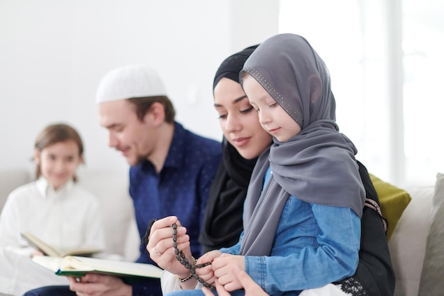 Tradycyjni muzułmańscy rodzice rodzinni z dziećmi czytającymi Koran i modlącymi się razem na kanapie przed kolacją iftar podczas uczty ramadanu w domu