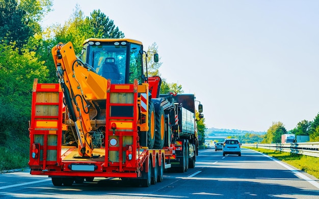 Transporter samochodowy z ciągnikami w drodze. Niski holownik pojazdów samochodowych na podjeździe. Europejska logistyka transportu w transporcie prac przewozowych. Przyczepa do transportu ciężkiego na autostradzie.