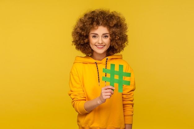 Trendy internetowe. Portret szczęśliwej kobiety z kręconymi włosami w bluzie z kapturem w miejskim stylu z dużym hashtagiem, co oznacza rekomendację bloga, tagowanie wirusowego posta. strzał w studio na białym tle na żółtym tle