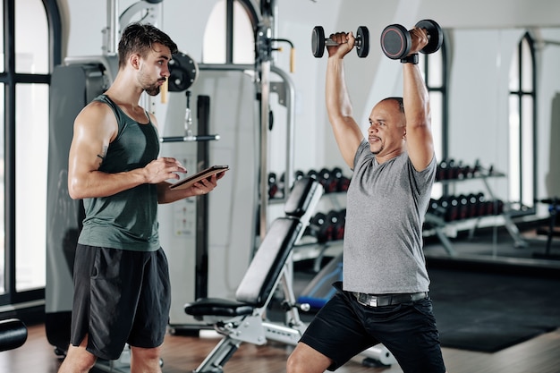 Trener fitness robienie notatek w dokumencie na komputerze typu tablet, gdy jego klient podnosi ręce z ciężkimi hantlami i robi rzuty