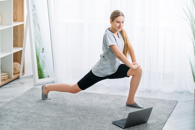 Trening fitness online Dziecko sport domowy Trening w pomieszczeniu Aktywny styl życia Wysportowana dziewczyna w stroju sportowym robi ćwiczenie lonżowania samotnie z laptopem w jasnym wnętrzu pokoju